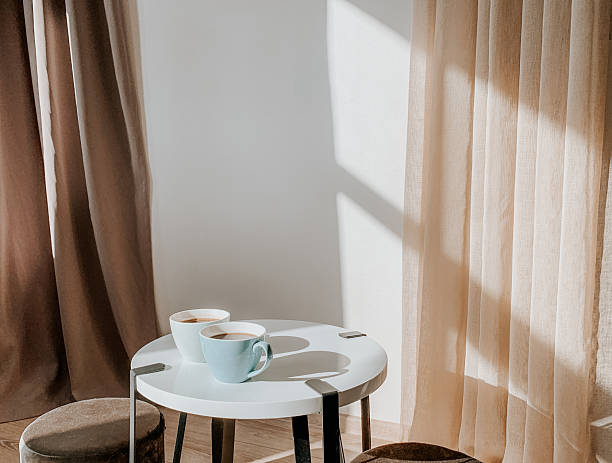 An inviting still life photograph showcases two ceramic cups filled with coffee or tea, resting on a minimalist white circular table. The scene is bathed in soft, natural light, casting gentle shadows on the wall behind. Elegant, semi-transparent curtains in calming beige and taupe hues drape on either side, adding a touch of warmth and texture to the cozy interior setting. This image evokes relaxation and comfort, ideal for lifestyle blogs, cafe promotions, or domestic-themed content.
