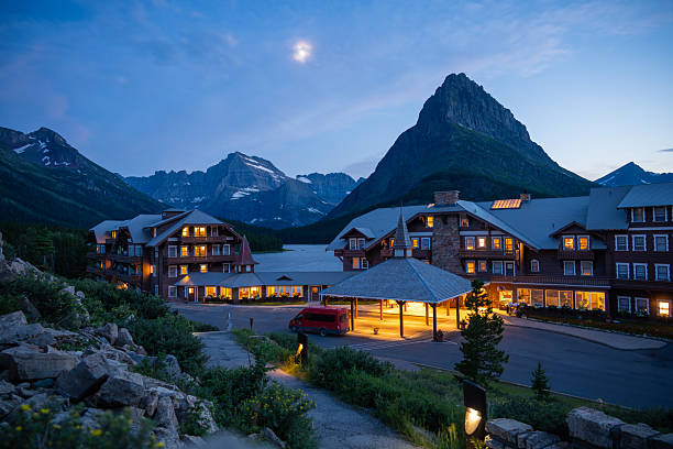 Many Glacier Hotel illuminated at dusk with the moon visible above dramatic alpine peaks in Glacier National Park, Montana, USA. Iconic mountain lodge during blue hour featuring warm lights, rugged Rocky Mountain scenery, and tranquil wilderness atmosphere, ideal for travel, tourism, hospitality, vacation, and destination concepts.