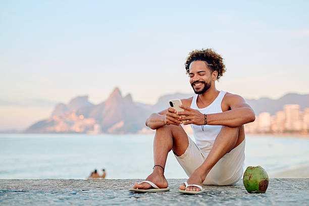 Brazilian man relaxing on a wall near Ipanema beach, using a cell phone and enjoying a fresh coconut drink