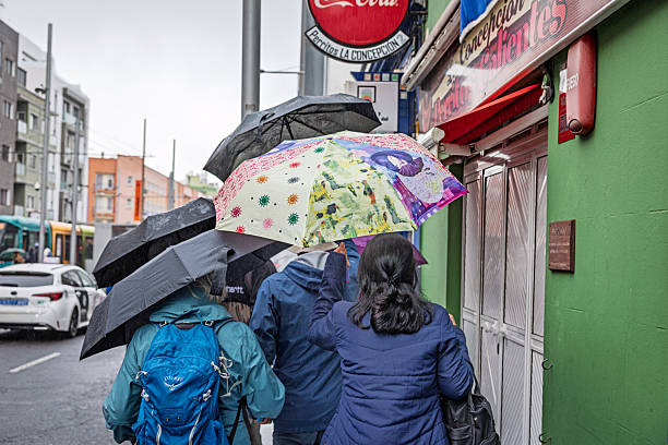 San Cristobal de La Laguna, Tenerife, Spain, Europe - December 18th 2025: Four people walk with umbrellas along a narrow, rain‑soaked sidewalk in San Cristóbal de La Laguna, the former capital of Tenerife in the Canary Islands. The historic city, known for its preserved colonial layout and UNESCO‑listed urban centre, attracts many visitors even in wet weather. The scene shows everyday pedestrian movement in one of the island’s most culturally significant towns.