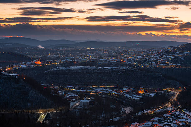 High angle view of the illuminated medieval Tsarevets Fortress and the traditional architecture of Veliko Tarnovo, Bulgaria, during the blue hour in winter. Light trails from cars and snowy hills create a magical atmosphere.