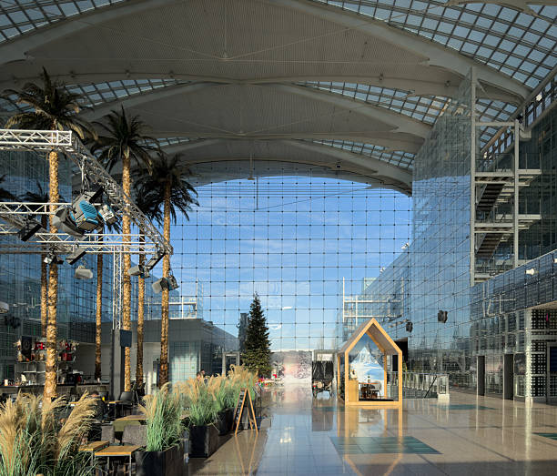 Munich, Germany - Dec 19 2025: Spacious modern atrium with a high glass ceiling and reflective floor, featuring indoor palm trees, seating areas, and a festive winter installation. A small wooden cabin with a winter scene backdrop and a decorated Christmas tree creates a seasonal contrast within the contemporary architectural space.