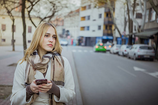 Beautiful young businesswoman is using a rideshare app on her mobile phone while waiting for a ride on the street on cold winter day. Female tourist waiting taxi ride for exploring new city.