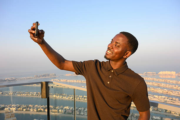 Young adult man enjoying his vacation, smiling while taking a selfie with his smartphone from a high rooftop overlooking the Palm Jumeirah in Dubai during sunset