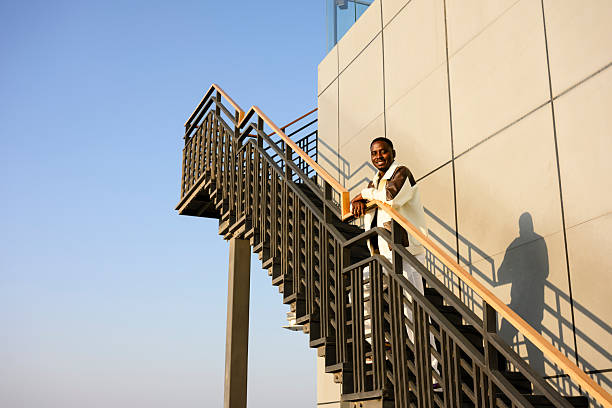 Caucasian young adult woman standing on outdoor steps playing acoustic guitar, smiling and looking upward, modern glass building and blue sky in background, casual urban scene