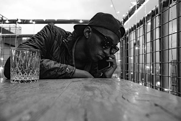 Black and white candid of a young man wearing a cap and sunglasses, holding a smartphone to his ear while leaning on a wooden table on a rooftop patio at night.