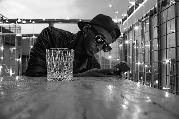 A black and white, medium-shot of a young man wearing a baseball cap and sunglasses while using a smartphone. He is seated at a wooden table on a rooftop balcony decorated with string lights. A crystal glass is visible in the foreground.