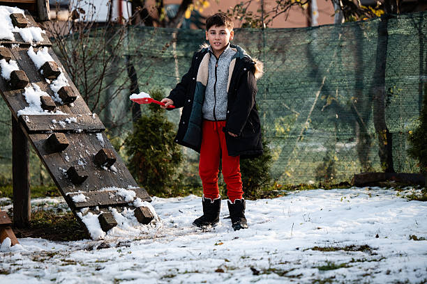 Boy standing in a snowy garden holding a red shovel, dressed in winter clothes, enjoying cold weather activities