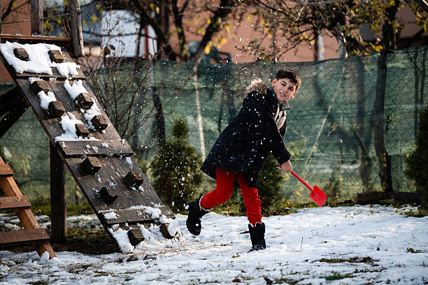 A young Caucasian girl bundled up in warm clothing smiles to the camera while sitting outside in the snow with a large shovel.