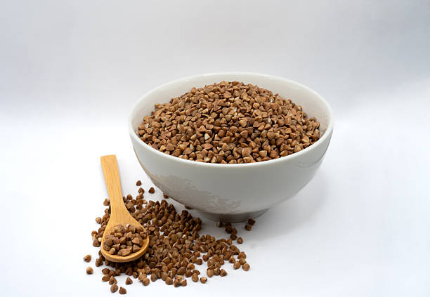 Raw buckwheat groats in ceramic bowl with scattered grains and wooden spoon. Natural healthy food concept with clean minimalist styling.