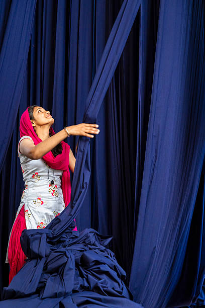 A woman in a white floral kurta and bright pink dupatta reaches up to pull or arrange long, deep-blue fabric panels. She looks upward with a focused, gentle expression while a large pile of the same navy material rests at her feet. The scene is dominated by vertical swathes of rich blue cloth, creating strong color contrast with her pink scarf and embroidered outfit.