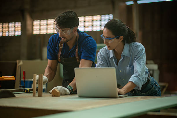 Carpenter with supervisor works on measuring and adjusting wooden piece on  workbench while checks project details on laptop craftsmanship with digital technology planning