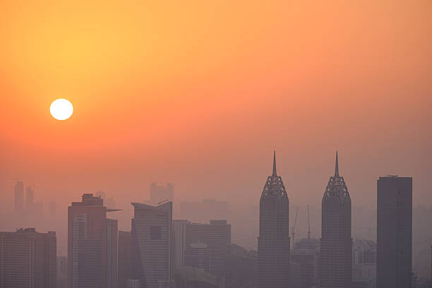 Dubai skyline silhouette of modern skyscrapers rising through haze and fog under a vibrant orange sunset, dramatic warm golden-hour cityscape over the desert metropolis