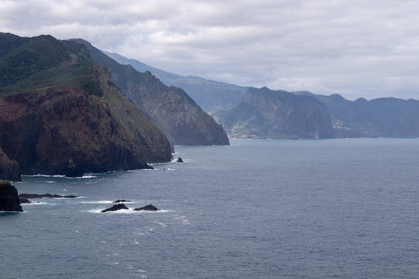 View of the rugged Atlantic coastline of Madeira Island, Portugal, with layered volcanic cliffs and distant headlands fading into the horizon. The calm ocean and overcast sky create a moody coastal landscape that highlights the island’s dramatic terrain and natural depth.