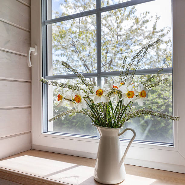Vertical spring still life with daffodils in white ceramic jug on wooden windowsill, bright window with mosquito screen, Scandinavian cabin interior, natural daylight, calm home decor mood
