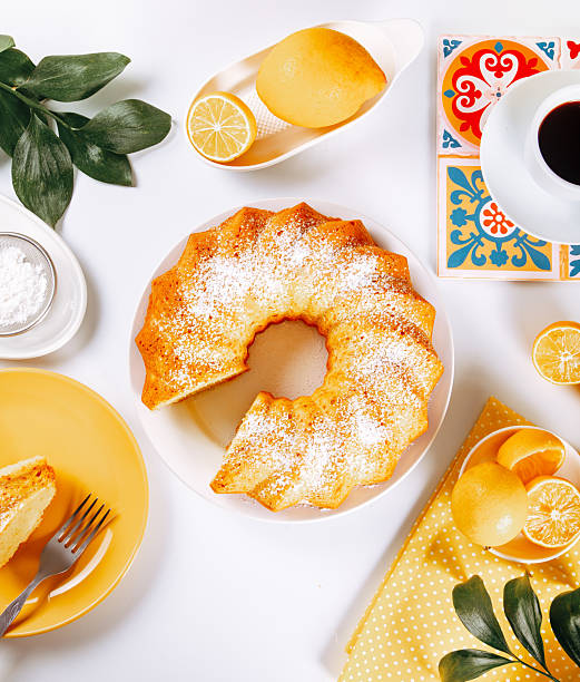 A freshly baked lemon bundt cake dusted with powdered sugar, served on a white plate with sliced lemons, a cup of coffee, and vibrant tableware on a cozy, bright background.