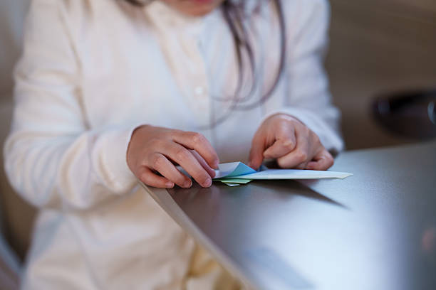 Close-up of a young child’s hands folding a paper airplane on a table inside a private jet cabin. The scene focuses on the simple activity during a flight, with soft natural light and aircraft interior details visible in the background.