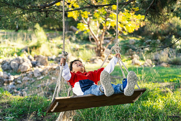 Smiling young boy enjoying a wooden swing hanging from a tree in a green outdoor setting. Childhood happiness, playtime, and carefree moments in nature.