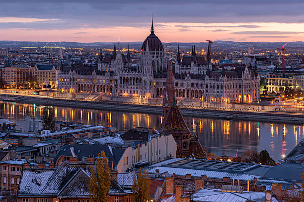 Panoramic view of the Hungarian Parliament Building and the Danube River at dusk in Budapest, Hungary. The image shows the illuminated neo-Gothic landmark with its golden lights reflecting on the water, framed by the snow-dusted rooftops of the Buda district and the spire of the Calvinist Church in the foreground.