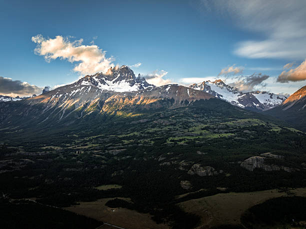 Expansive view across a green valley with forest patches and distant snow-covered peaks. The composition highlights the transition from fertile lowlands to rugged mountain terrain.