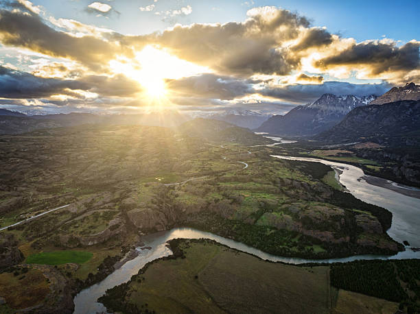 Dramatic landscape view with sunlight breaking through clouds and illuminating a winding river and open valley. The interplay of light and shadow creates a powerful sense of atmosphere and depth.