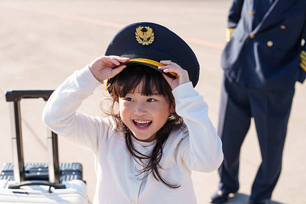 A happy young girl tries on a pilot’s cap and smiles confidently on an airport runway near a private jet, while her family and cabin crew stand in the background. The scene captures a joyful family travel moment, combining childhood imagination, luxury aviation, and premium customer experience in a bright outdoor setting.