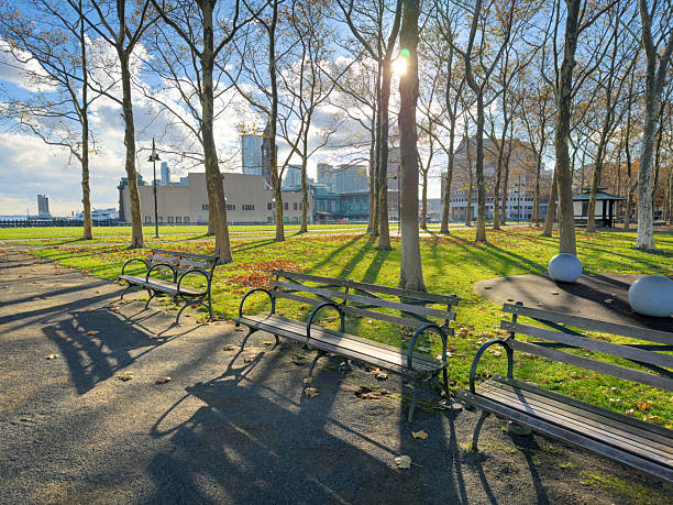 Wide angle view of several empty benches along a paved path in a New Jersey riverside park, with bare trees, bright winter sun casting long shadows, and city buildings visible in the background. This full frame outdoor scene is ideal for concepts of solitude, relaxation, urban nature, and seasonal change in editorial or commercial projects.