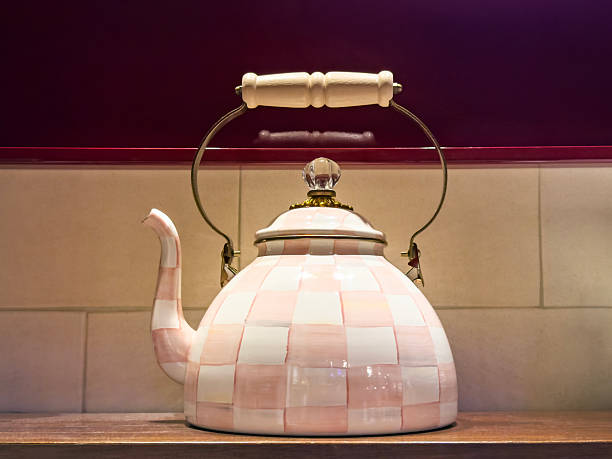 Still life photograph of a vintage enamel kettle with a curved spout and metal handle placed on a kitchen shelf against a tiled backsplash. Warm ambient lighting and soft shadows highlight the pastel tones and rounded shape of the kettle, creating a cozy home kitchen, rustic decor, and nostalgic cooking concept with ample copy space for text and design elements.