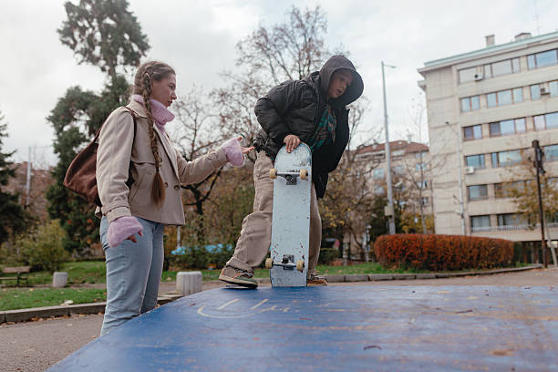 Low angle child lifting board nose, woman nearby ready support, cloudy sky, apartment blocks background, fall leaves ground, beginner skateboarding moment