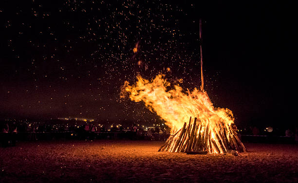 Large bonfire arranged in a pyramid shape on the sandy beach at night, flames rising and sending sparks into the air. The warm glow contrasts with the dark surroundings, creating a dramatic and atmospheric scene. Perfect for illustrating outdoor activities, camping, beach gatherings, firelight ambiance, or nighttime events.