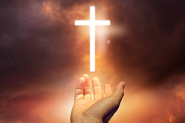 A human hand reaches upward toward a glowing white cross against a dramatic sunset sky, symbolizing faith, hope, and salvation.