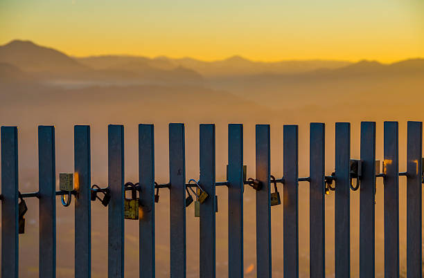 Close-up of a metal fence at a scenic viewpoint, covered with love locks placed by couples. The warm glow of the sunset illuminates the mountains in the background, creating a romantic and picturesque atmosphere. Ideal for themes of romance, travel, relationships, tourism, and scenic landscapes.