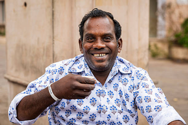 A warm, candid portrait of a middle-aged man smiling directly at the camera. He wears a white shirt patterned with small blue floral motifs and a simple wristband, and holds a small cup near his mouth as if drinking tea. The neutral stone background suggests an outdoor setting; his bright, open smile and relaxed posture convey friendliness and cheerfulness.