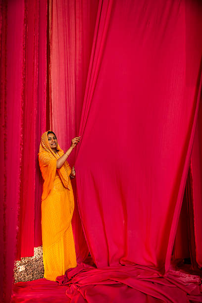 A smiling woman dressed in a bright yellow sari and headscarf stands amid towering panels of vivid magenta-red fabric, pulling one curtain aside. The composition emphasizes the flowing textures and saturated colors as the cloth hangs and pools on the ground; small pebbles are visible at her feet. Warm light highlights the folds, the woman's bangles, and the rich contrast between her clothing and the surrounding fabrics.