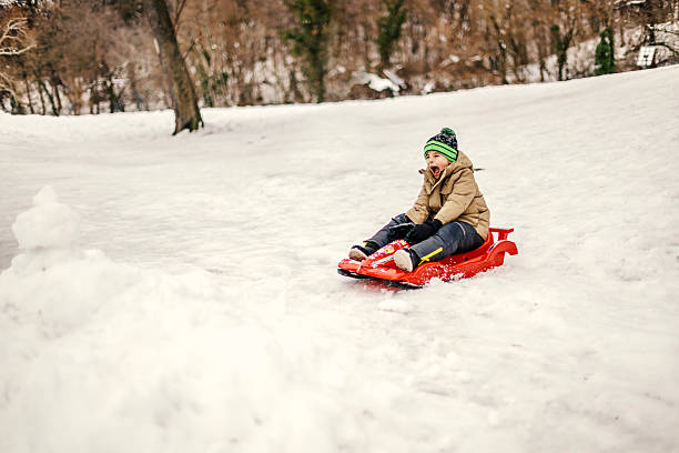 Young boy laughing as he sleds down a snowy hill on a red plastic sled, bundled in winter gear and enjoying active outdoor play and joyful cold-weather fun