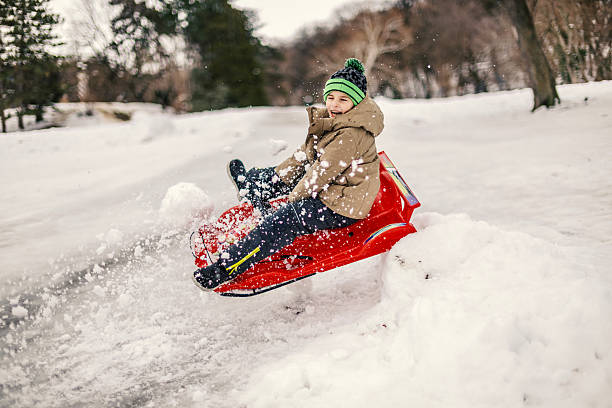 Young boy experiencing pure joy and excitement as he speeds down a snow-covered slope on his red sled, creating a flurry of snow around him during a fun outdoor winter activity