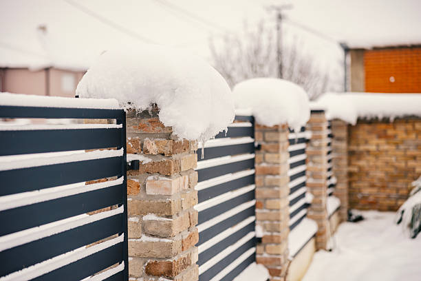 Contemporary black metal and brick fence posts are covered in a thick layer of fresh white snow and icicles on a clear, cold winter day with a residential building visible in the background