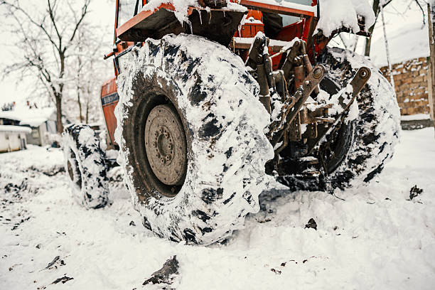Heavy-duty orange farming tractor standing idle in a rural environment during a cold snowy winter day, its large tires covered with fresh snow and ice