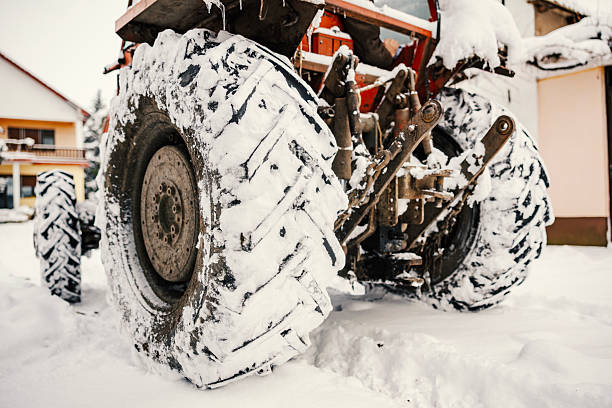 Farm tractor standing idle outdoors during heavy winter snowfall, its large tire treads filled with fresh snow and icicles hanging from the machine, creating a challenging rural weather scene