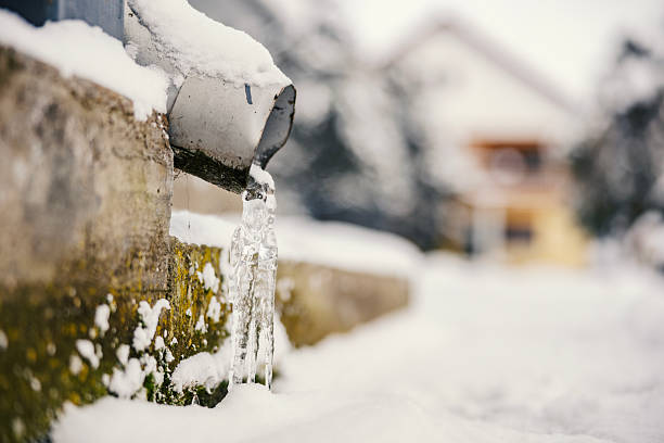 Drainpipe shedding water and forming a long icicle, surrounded by white snow and concrete covered in green moss, with a blurred house in the background