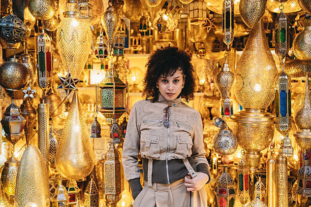 Young adult woman standing among a vibrant display of golden metallic lanterns in a traditional Moroccan souk market