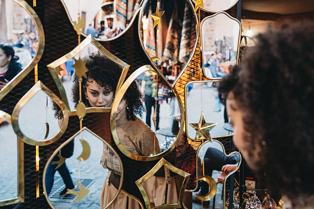 Young adult woman with curly hair looking at her reflection in ornamental brass mirrors at a busy Marrakech market