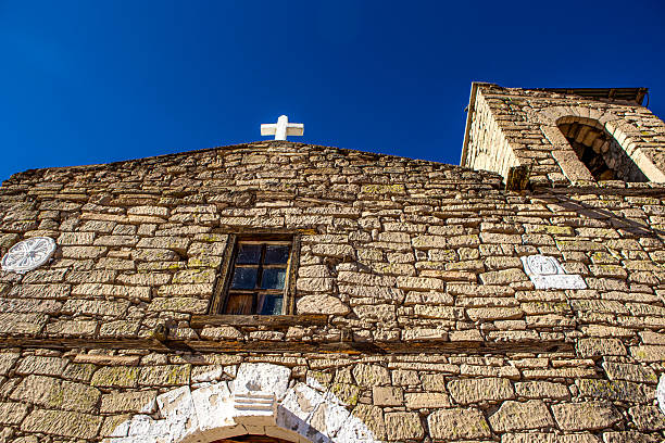 This photograph shows a small historic stone chapel located in a rural mountain village in northern Mexico, likely within the Sierra Tarahumara region of Chihuahua. The simple colonial-style architecture, bell tower, and wooden door reflect traditional Catholic heritage in remote communities, surrounded by rocky landscapes and highland terrain.