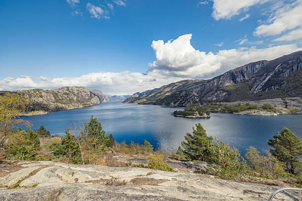 Wide panoramic view of Lysefjord in Norway, showing calm blue fjord waters framed by steep rocky mountains, forested slopes, and small islands. Classic Norwegian fjord landscape captured from an elevated viewpoint in daylight under partly cloudy sky.