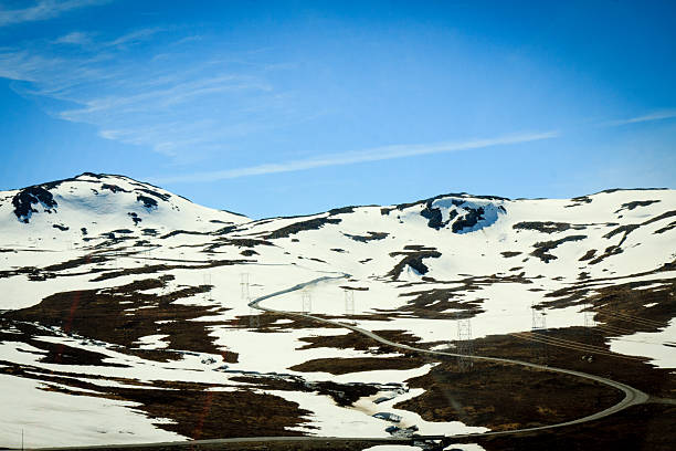 A high-altitude winding road cuts through Jotunheimen National Park on a sunny day, surrounded by snow-covered mountain terrain.