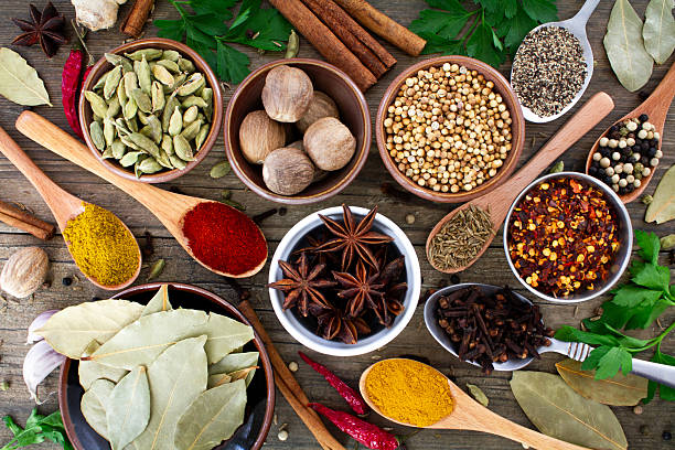 A selection of healthy cooking ingredients including dried spices in spoons and bowls shot on a dark wood background. Cloves, cardamom, cumin seeds, chilli powder, turmeric, thyme and peppercorns.With fresh chilli peppers, cinnamon, fresh onions, garlic, cilantro and nutmeg. Shot from above.