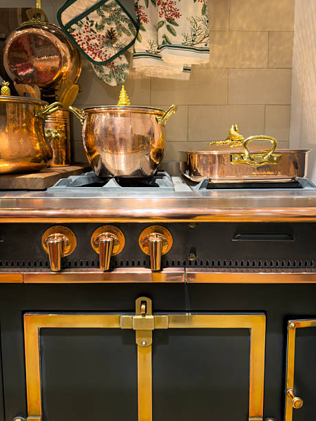 Vertical still life photograph of a dark rustic stove topped with shiny copper pots, pans, and kettles in a warm domestic kitchen. The golden metal surfaces, vintage stove details, and soft ambient lighting create a nostalgic farmhouse cooking, traditional cuisine, and cozy home interior concept with ample copy space for text.