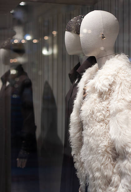 Shop window with two female mannequins with silhouettes instead of faces. One mannequin is wearing a white fur coat, the other a black shirt and a black hat with glitter. The window reflects the glare from the lamps and garlands.