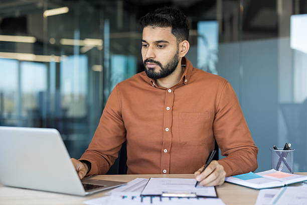 istock Indian man working on laptop and writing notes in modern office 2256100092