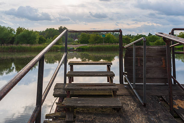 Close-up view of old wooden steps and metal railings on a small dock above calm lake water. Rustic textures, soft reflections, and peaceful shoreline scenery create a mood of solitude, slow travel, and nature escape.